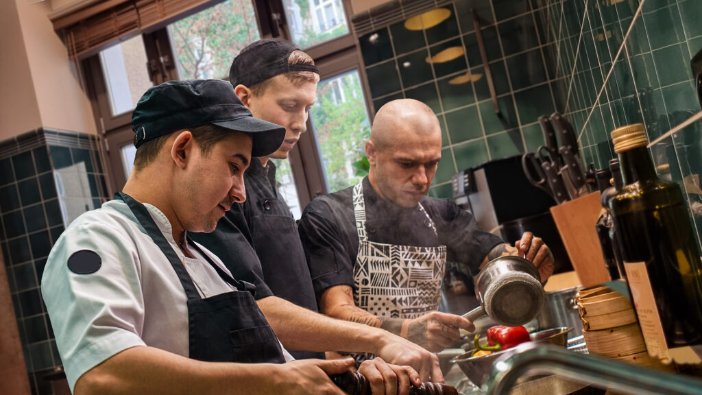 Teamarbeit. Küchenchef und seine beiden Assistentinnen in Schürzen kochen ein neues Gericht in einer modernen Küche, Kochvorgang