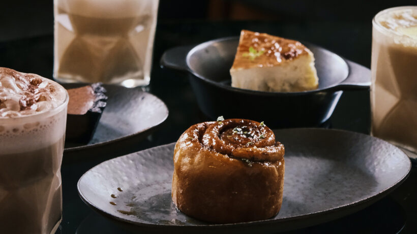 drei Kaffeegläser mit Milchschaum und zwei Stück Kuchen und Gebäck angerichtet auf dunklen Keramiktellern