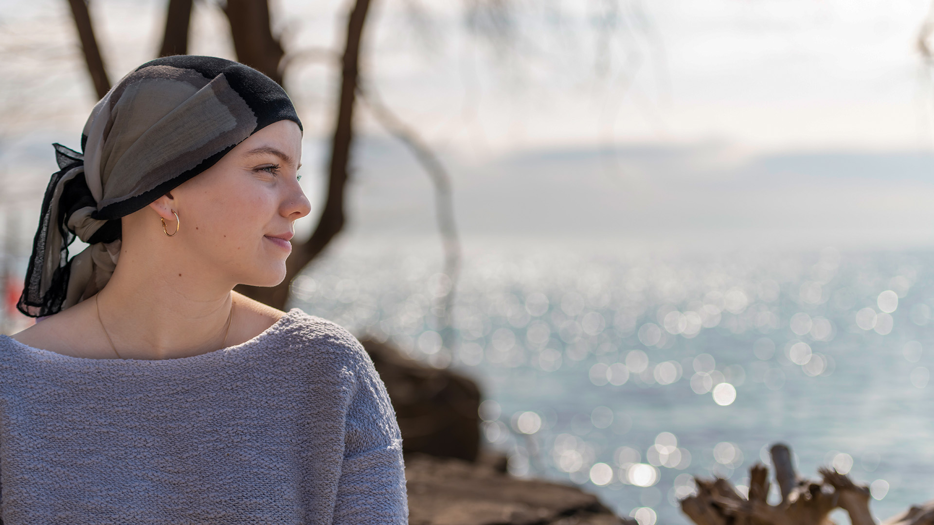 Eine junge krebskranke Frau lächelt selbstbewusst und schaut in die Ferne. Sie trägt ein Kopftuch und entspannt sich im Freien. Im Hintergrund sind ein See und ein Baum zu sehen. Es ist ein sonniger, friedlicher Tag.